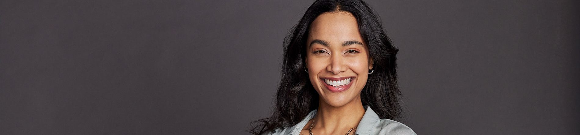 The image is a photograph of a woman with light skin, smiling at the camera. She appears to be in her late twenties or early thirties and has long hair. Her eyes are looking directly at the camera, and she is holding up her index finger near her mouth as if she s making a point or emphasizing something. The background is plain and light-colored, which suggests that this could be a stock photo used for various purposes such as advertising, personal branding, or lifestyle content.