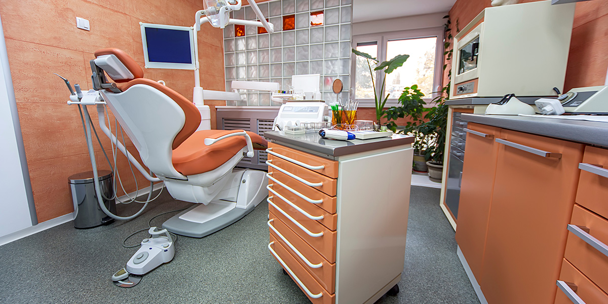 The image shows an interior view of a dental office with a modern chair and equipment, including a computer screen and a counter with drawers.