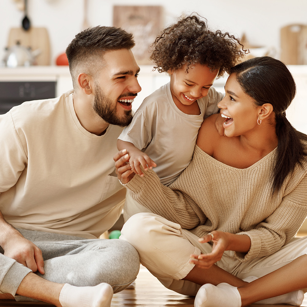 This is a photograph showing a family of four - a man, a woman, and two children - sitting on the floor indoors, smiling and looking at the camera. The adults appear to be young adults, while one child is a young boy and the other is an infant girl. They are all dressed in casual clothing, and the setting seems warm and cozy with a homely atmosphere.