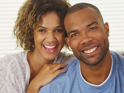 The image depicts two individuals, likely a couple, posing closely together with smiles on their faces, both looking directly at the camera.